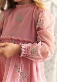 Enfant portant une robe rose à manches longues avec des manches florales brodées transparentes et des détails aux poignets.