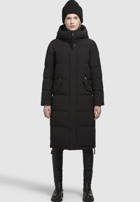 Kvinde iført en lang sort polstret vinterfrakke med hætte, sort hue, sorte bukser og sorte støvler, der står op ad en ensfarvet baggrund.