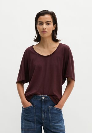 Jeune femme aux cheveux courts et foncés porte un t-shirt bordeaux ample et un jean bleu, les mains dans les poches, debout devant un fond uni.