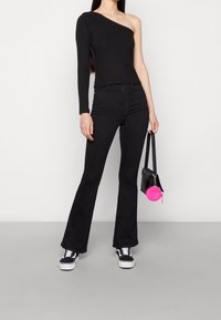 Top negro de canalé con un solo hombro combinado con jeans negros acampanados. La modelo sostiene un pequeño bolso negro con un detalle circular rosa. Zapatillas negras.