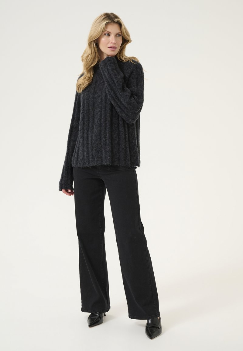 Dunkelgrauer Strickpullover mit vertikaler Rippung, lockerer Passform, hohem Kragen und langen Ärmeln, kombiniert mit schwarzen, weit geschnittenen Hosen und spitzen Schuhen.