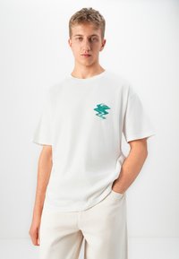 Ung mand iført hvid t-shirt med grønt abstrakt logo på brystet, poserer med den ene hånd i lommen mod en ensfarvet hvid baggrund.