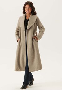 Beige Wollmischungsjacke mit großem Schalkragen, doppelten Knöpfen und seitlichen Taschen, kombiniert mit dunklen Jeans und schwarzen Stiefeln mit Absatz.