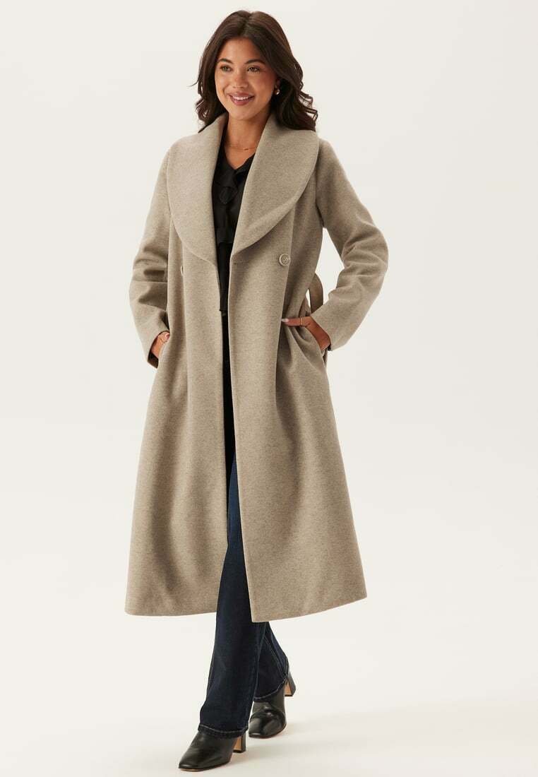 Beige Wollmischungsjacke mit großem Schalkragen, doppelten Knöpfen und seitlichen Taschen, kombiniert mit dunklen Jeans und schwarzen Stiefeln mit Absatz.