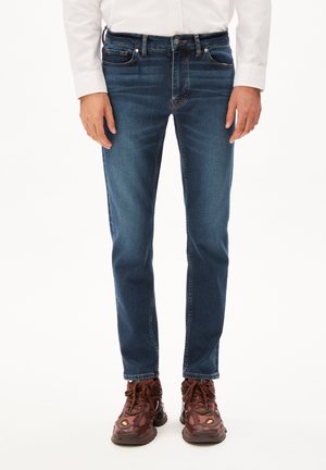 Jeans oscuros de corte slim con detalles desvaídos, diseño de cinco bolsillos y costuras en contraste, combinados con zapatillas marrones.