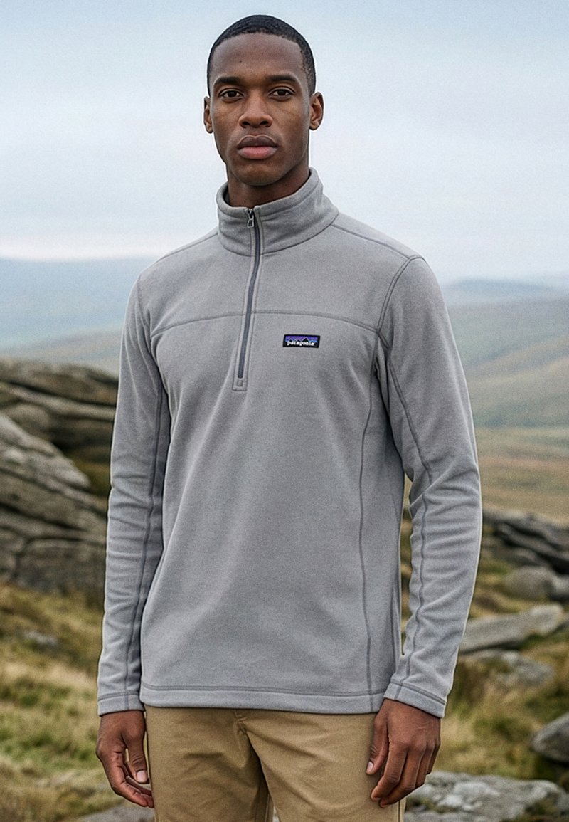Mann trägt hellgrauen Patagonia-Quarter-Zip-Fleece und beigen Hosen, steht im Freien vor einer felsigen Landschaft.