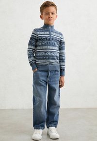 Plavi pleteni pulover s bijelim uzorcima, s patentnim zatvaračem; uparen s širokim plavim trapericama i bijelim tenisicama.