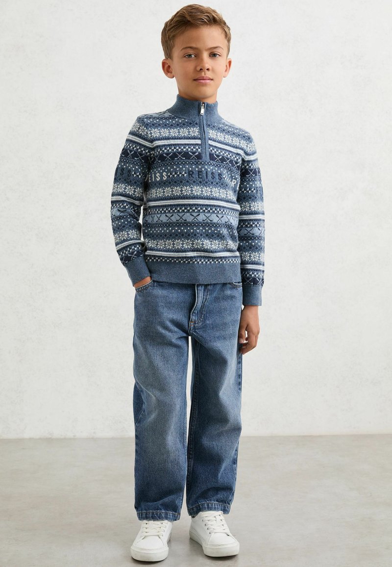 Plavi pleteni pulover s bijelim uzorcima, s patentnim zatvaračem; uparen s širokim plavim trapericama i bijelim tenisicama.