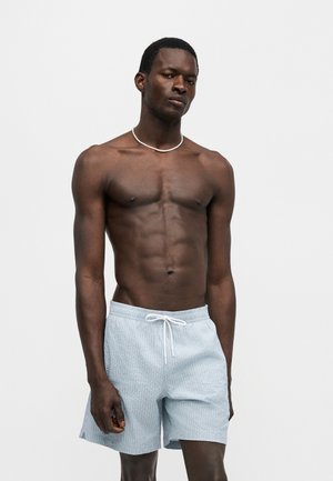 Homme musclé à la peau foncée portant un short bleu clair texturé avec cordon de serrage et un collier blanc, debout devant un fond blanc uni.