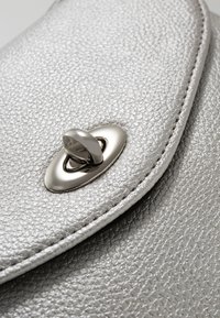 Primo piano di una borsa in pelle argentata con texture, caratterizzata da una chiusura ovale metallica con una forma distintiva e dettagli particolari.