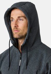 Mörkgrå zip-up hoodie med snörhuva. Har en mjuk textur och svart dragkedja, med en märkesetikett nära dragkedjan.