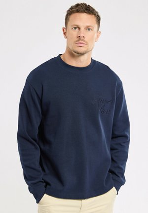 Homme aux cheveux courts portant une chemise bleu marine à manches longues avec un texte brodé sur la poitrine, mains dans les poches, fond neutre.