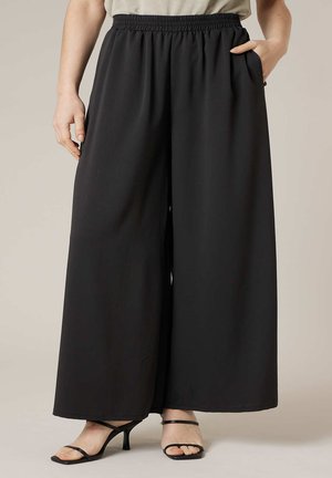 Femme portant un pantalon large noir avec taille élastique et des sandales à talons noires à brides, main dans la poche.