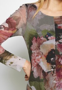 Gros plan d'une personne portant un haut transparent à manches longues avec un motif floral multicolore, la main reposant sur la hanche.