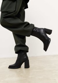 Bottes de cheville noires à talons en cuir lisse avec un talon carré épais et une semelle texturée, associées à un pantalon vert à jambes fuselées.