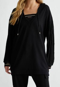 Schwarzer Kapuzenpullover mit V-Ausschnitt und Kordelzügen. Mit strukturierten Spitzeärmeln und goldenen Akzenten an den Bündchen. Lässige Passform mit einem geschichteten Look.