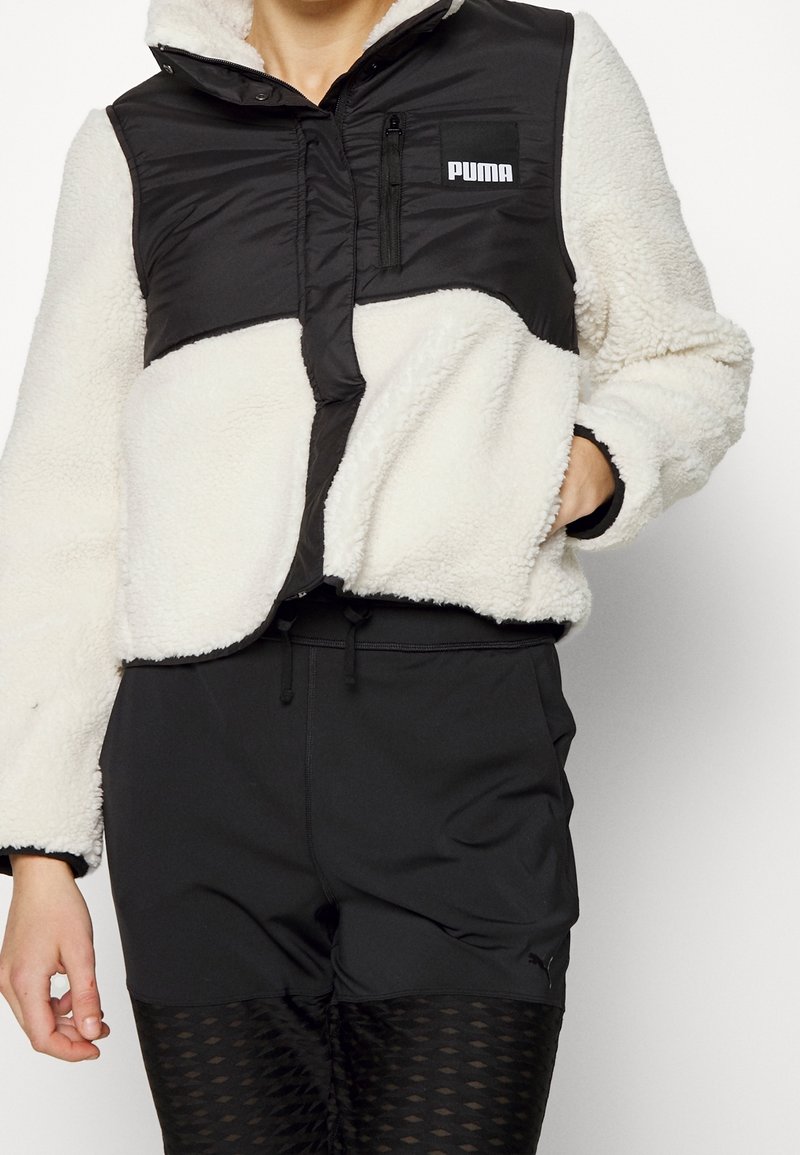 Veste en polaire blanche avec panneau noir sur la poitrine et logo Puma, portée avec un short noir et des leggings noirs à motifs.