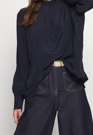 Kobieta w luźnej granatowej bluzce w delikatne kropki, włożonej do wysokich dżinsów z ciemnego denimu z wyraźnym białym przeszyciem i beżowym paskiem.