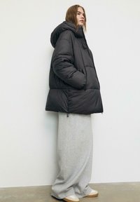 Giubbotto di piumino nero con un alto colletto e cappuccio, caratterizzato da una forma squadrata, grandi sezioni trapuntate e tasche laterali, indossato sopra pantaloni grigi.