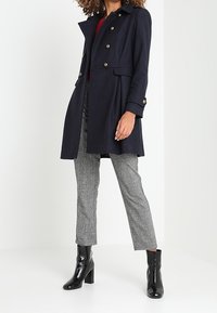 Cappotto in misto lana blu navy con design doppiopetto, bottoni dorati e colletto alla moda. Abbinato a pantaloni grigi a quadri e stivaletti neri lucidi.