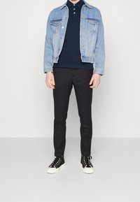 Džínová bunda se světle modrým omyvem, tmavé polo tričko, černé slim-fit kalhoty a černé tenisky s bílými podrážkami a šněrováním.