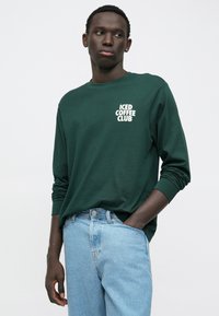 Tamna zeleni majica dugih rukava s bijelim logotipom "ICED COFFEE CLUB". Uparen s svijetlosivim trapericama. Pamuk, opušteni kroj.