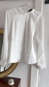 White long-sleeve blouse with a high neckline, pleated front detail, and buttoned cuffs. Made of smooth, lightweight fabric.