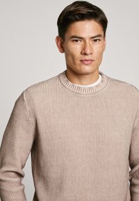 Vaalea beige ribattupaita, jossa on pyöreä pääntie. Edessä on tekstitetty neulekuvio ja tiukat hihat. Paita on puettu valkoisen paidan päälle.