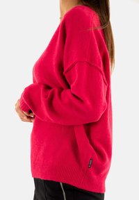 Fuchsia oversized trui met geribbelde manchetten en onderrand, met een zijsplit en een klein merklabel aan de zijkant. Zachte, gestructureerde stof.