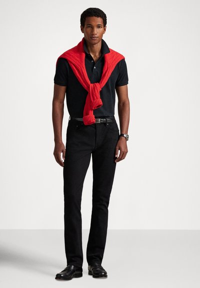 Melns polo krekls ar salocītu apkakli, sarkans adīts džemperis pār pleciem, melnas džinsa bikses un melni ādas apavi, viss uz vienkārša fona.