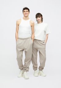 Δύο μοντέλα στέκονται δίπλα δίπλα, φορώντας βαμβακερό T-shirt και αμάνικη μπλούζα σε κρεμ απόχρωση, με μπεζ φόρμες και λευκά αθλητικά παπούτσια.