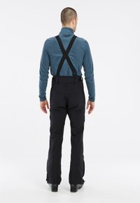 Zwarte skibroeken met verstelbare bretels, met een gladde textuur en zijzakken met rits, gedragen over een blauwe fleece top.