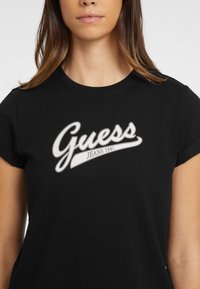 Czarna koszulka z krótkim rękawem wykonana z bawełny, z dużym białym logo "Guess Jeans 1981" w kursywie na przodzie.