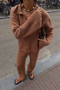 Rjav pleten dvo delni komplet, ki vključuje gumbasto jopico in usklajene široke hlače. Tekstura je rebrasta z ohlapnimi rokavi.