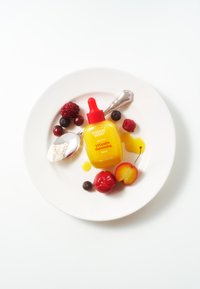 Serum witaminowe w żółtej butelce z czerwoną pipetą na białym talerzu. Wokół rozsypane świeże owoce oraz łyżeczka z proszkową substancją.