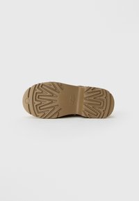 Semelle de chaussure marron avec des motifs de bande de roulement en zigzag et courbes et un petit logo en forme d'éclat de soleil près de la zone des orteils, sur un fond pâle.