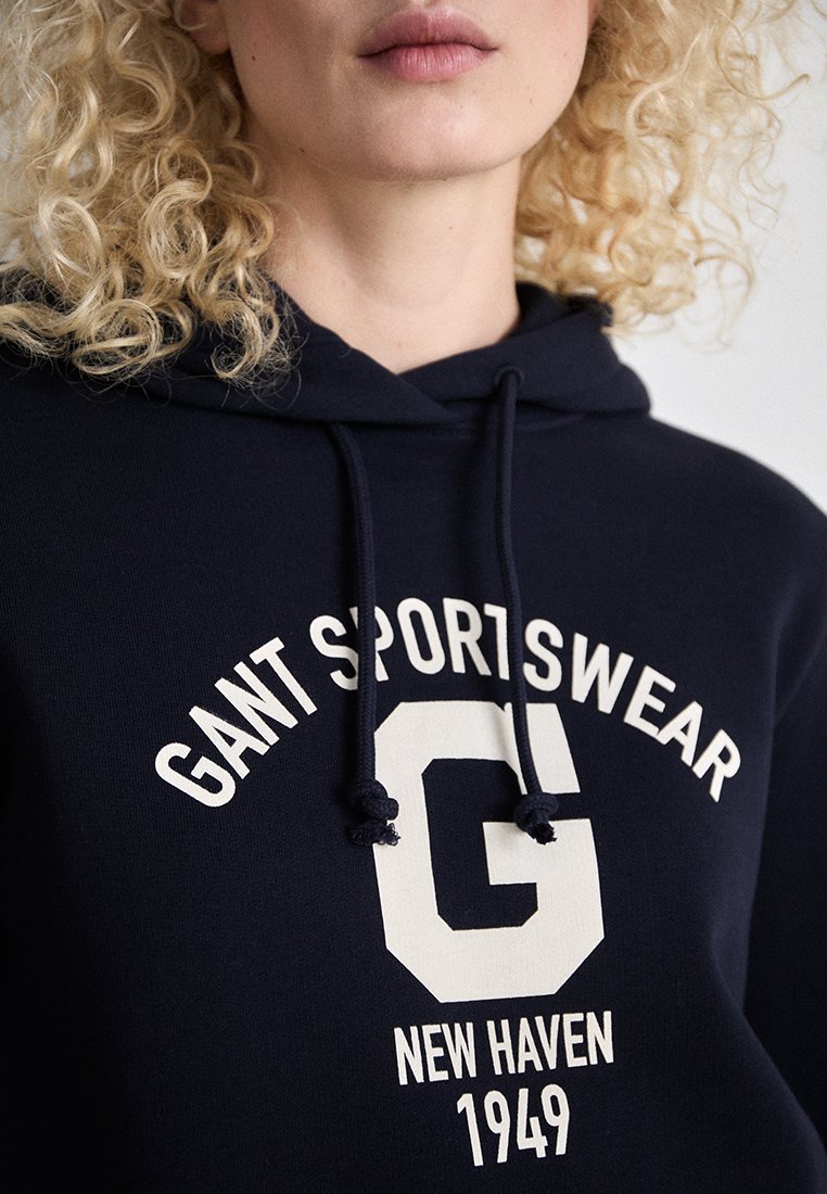 Navyblaues Hoodie mit weißem Text "GANT SPORTSWEAR". Es hat eine Kordelzugkapuze und einen großen grafischen Buchstaben "G" auf der Vorderseite.