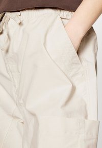 Beige utility shorts med snøre i taljen, sidelommer og en tekstureret stoffinish. Har en knælang kant og subtile syninger.