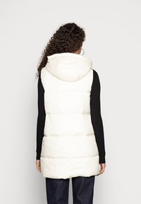 Leichter, cremfarbener Steppweste mit Kapuze, ausgestattet mit horizontalen Steppnähten und einem längeren Schnitt, kombiniert mit einem schwarzen Langarmoberteil.