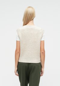 Bézs kötött ujjatlan pulóver kerek nyakkivágással, puha textúrával és enyhe csillogással; fehér ingre és olívazöld nadrágra viselve.