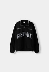 Svart sweatshirt med dragkedja vid kragen, med vita randiga detaljer, fet grå text "BUSHWICK" och ytterligare grafiska detaljer på framsidan.