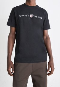Camiseta de algodón negra con el logo "GANT 1949" en blanco y emblema. Mangas cortas, corte estándar, cuello redondo, textura suave.
