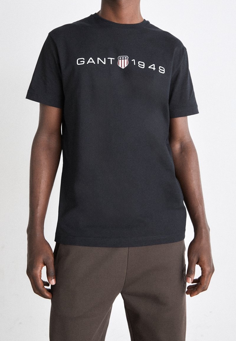 Camiseta de algodón negra con el logo "GANT 1949" en blanco y emblema. Mangas cortas, corte estándar, cuello redondo, textura suave.