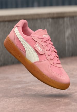 Różowy but sportowy Puma z białym paskiem bocznym i gumową podeszwą, pokazany pod kątem na rozmytym tle na zewnątrz.