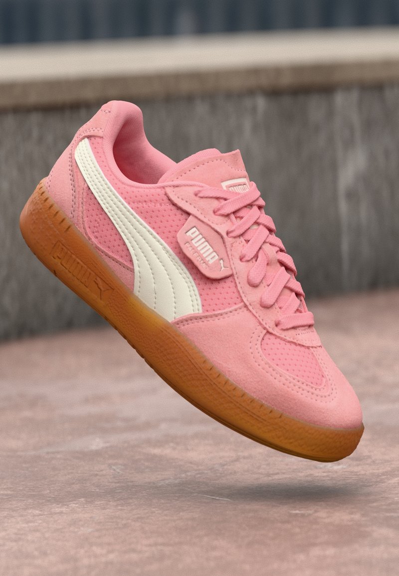Baskets roses Puma avec bande latérale blanche et semelle en gomme, montrées en angle sur un fond extérieur flou.