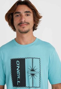 Camiseta de algodón turquesa con un diseño gráfico en negro que presenta un estallido central, bordeado por un patrón de cuadros y texto.