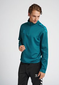 Sudadera con cremallera en color azul verdoso, con cuello alto y mangas largas, que presenta rayas negras en los hombros y un logo en el pecho.