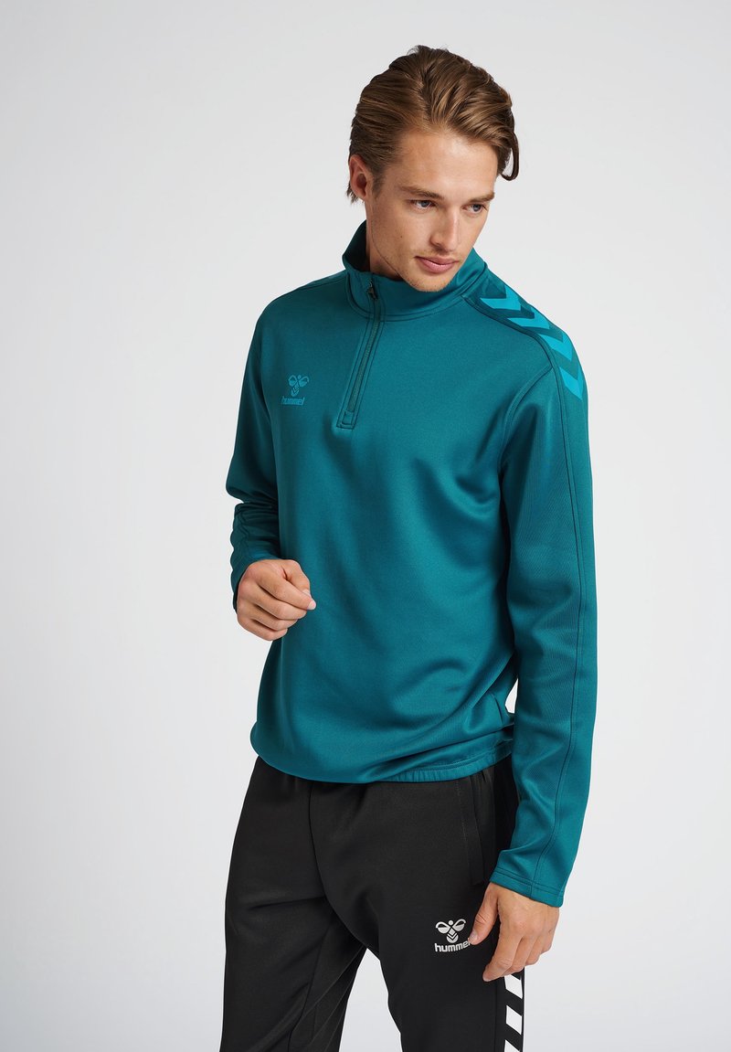 Sudadera con cremallera en color azul verdoso, con cuello alto y mangas largas, que presenta rayas negras en los hombros y un logo en el pecho.