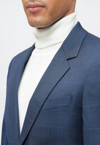 Σκούρο μπλε σακάκι με καρό σχέδιο, κατασκευασμένο από υφαντό ύφασμα, συνδυασμένο με λευκή ζιβάγκο. Διαθέτει μία τσέπη στο στήθος και καπόνια πέτο.