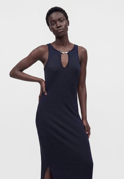 Femme portant une robe bleu marine sans manches avec une encolure en forme de trou de serrure devant et une fente latérale, debout avec une main sur la hanche.
